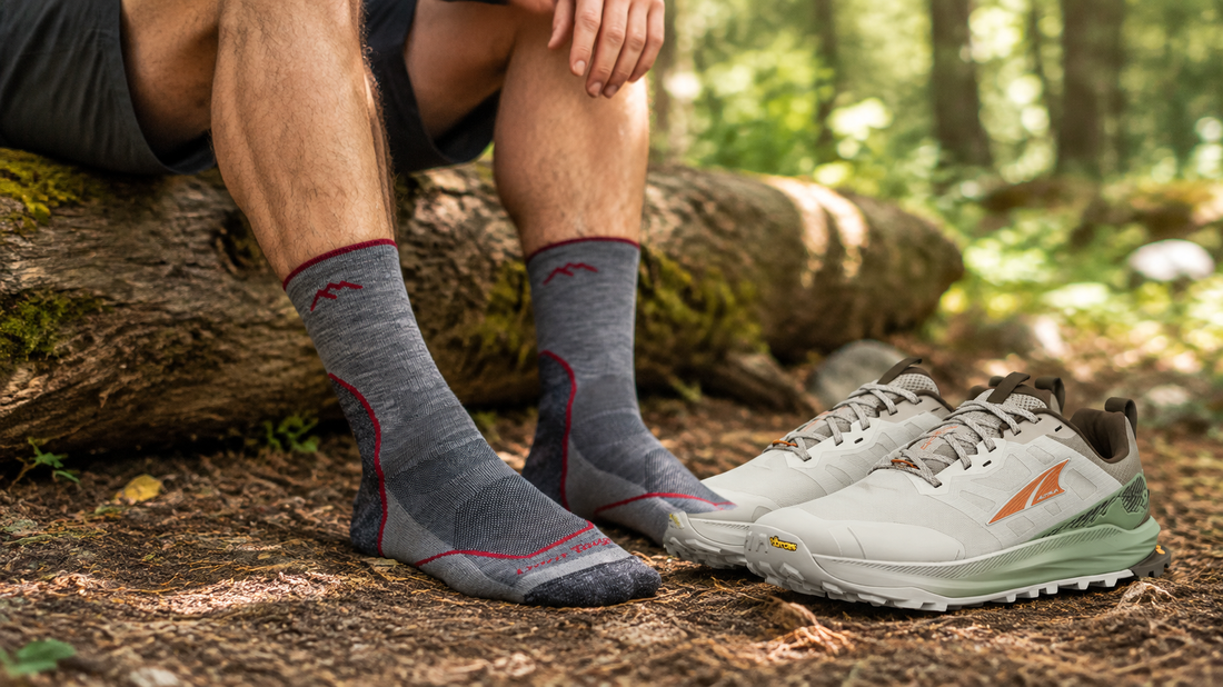 Hiker wears gray socks beside Altra trail shoes at a woodland rest stop