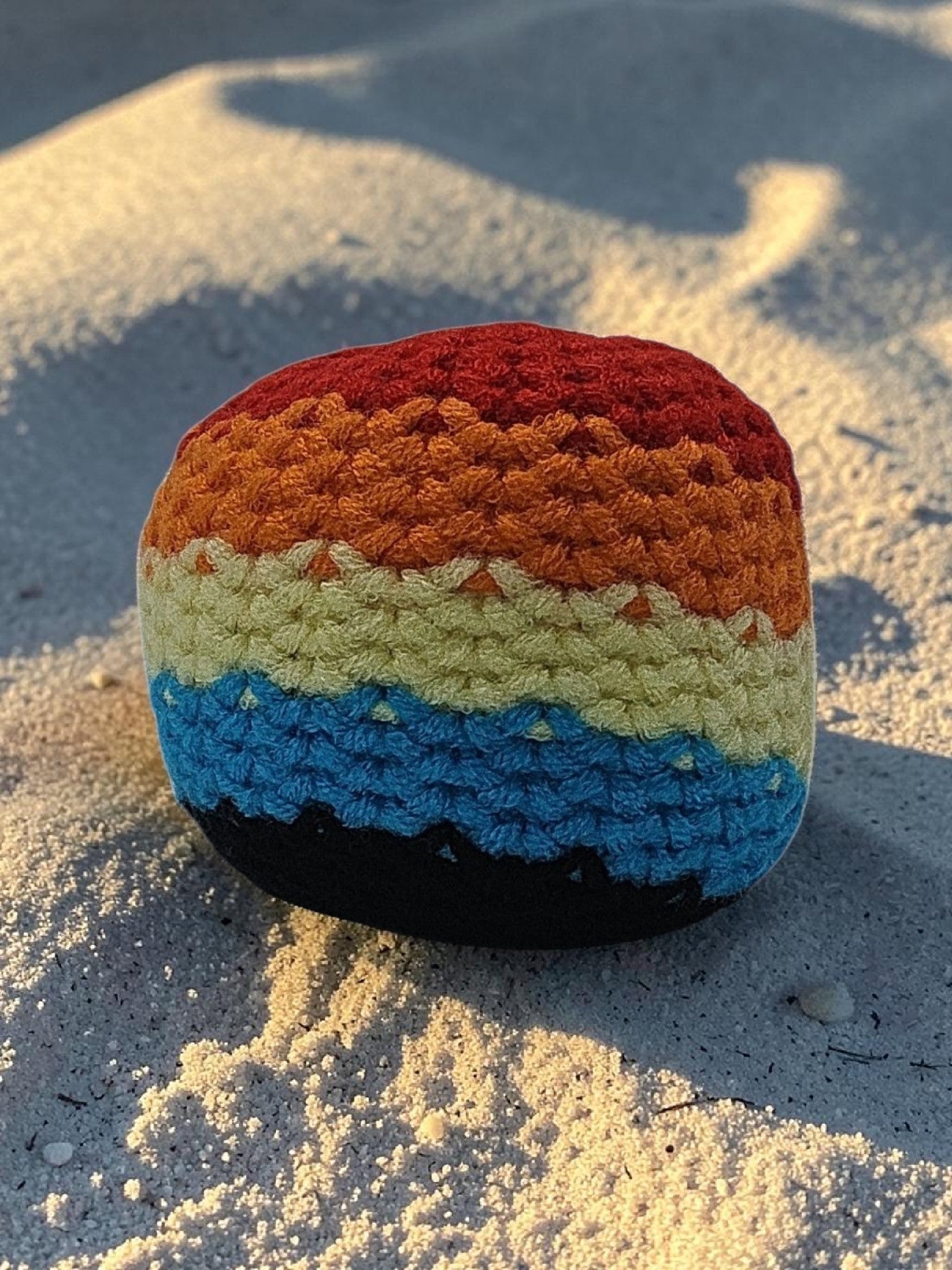 Colorful crocheted rainbow-patterned object on sandy ground